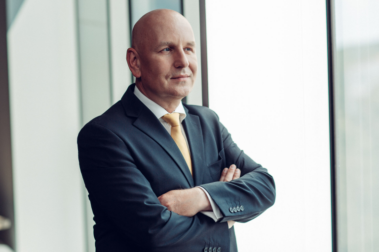 Biznesa portrets ar profesionāli ģērbtu vīrieti uzvalkā, stāvot pie lieliem biroja logiem ar sakrustotām rokām, skatoties tālumā. Attēls simbolizē līderību, profesionālismu un stratēģisku domāšanu.
